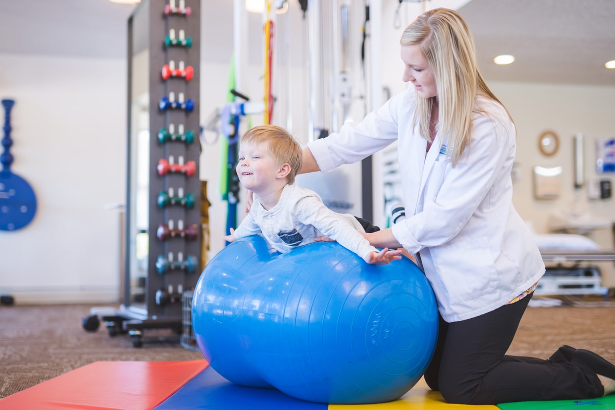 Young boy laying down on an exercise ball to improve balance alongside his physical therapist